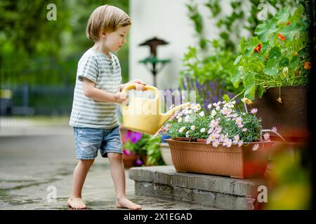 Süßer kleiner Junge, der am Sommertag im Hinterhof Blumenbeete tränkt. Kind mit Gießkanne zum Gießen von Pflanzen. Kind hilft bei den alltäglichen Aufgaben. Mommy Stockfoto