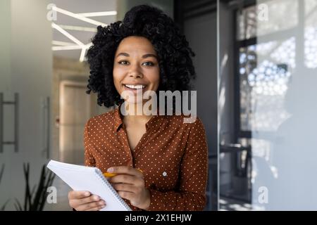 Eine fröhliche junge Frau mit lockigen Haaren nimmt an einem Online-Training Teil, macht Notizen und lächelt in einem modernen Büroumfeld. Stockfoto