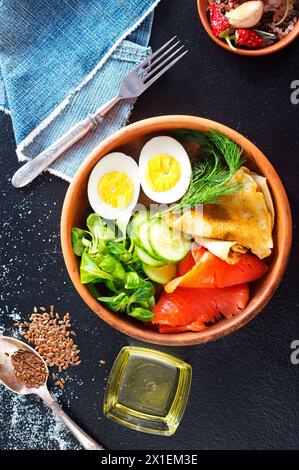 Köstlicher gesunder Brunch - gekochtes Ei, geräucherter Lachs, grüner Salat und herzhafte Pfannkuchen auf dunklem Hintergrund Stockfoto