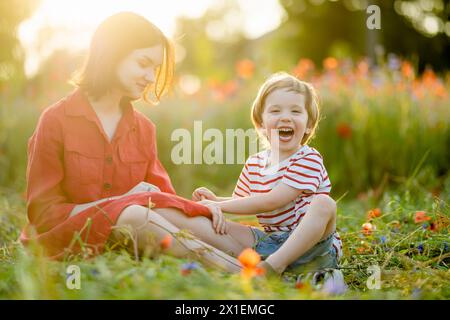 Wunderschönes Teenager-Mädchen und ihr kleiner Bruder bewundern Mohn- und Knabenkraut-Blumen in blühendem Mohnfeld an sonnigen Sommertagen. Stockfoto