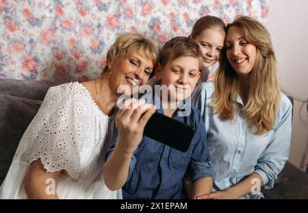 Eine vierköpfige Familie sitzt auf einer Couch, lächelt und posiert zusammen, während sie ein Selfie mit einem Smartphone macht. Stockfoto