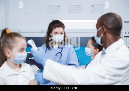 In einer Klinik untersucht schwarzer Arzt weibliches Kind und kommuniziert mit der Mutter über Gesundheit. Gesichtsmaske getragen, Thermometer verwendet, wo multirassisches medizinisches Personal betont Gesundheit und Medizin. Stockfoto