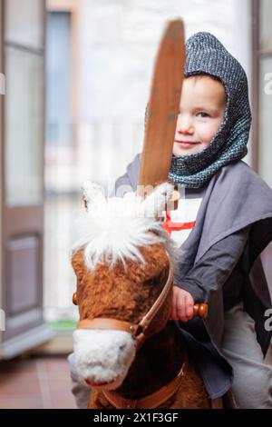 Junge als Ritter gekleidet, der auf seinem Schaukelpferd reitet und sein Spielzeug-Holzschwert zeigt, während er in die Kamera blickt. Stockfoto