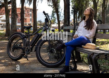 Die junge lateinamerikanische Frau (33) sitzt auf einer Bank auf dem Platz neben ihrem Elektrofahrrad. Konzeptmittel für Transport und Frühling. Stockfoto