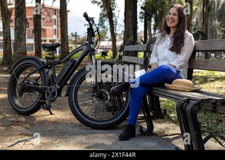 Die junge lateinamerikanische Frau (33) sitzt auf einer Bank auf dem Platz neben ihrem Elektrofahrrad. Konzeptmittel für Transport und Frühling. Stockfoto