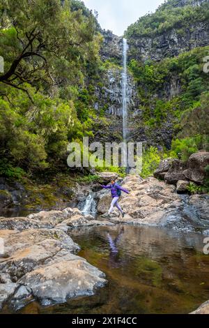 Beschreibung: Weibliche Touristenin mit Rucksack springt über Felsen am Wasserfall, der von einer hohen Felswand fällt. Lagoa do Vento Wasserfall, Madeira Insel, Portugal Stockfoto