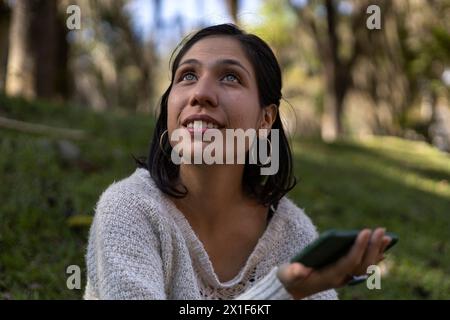 Junge lateinamerikanische Frau (29), die auf dem Gras sitzt und ihr Handy hält, während sie nach rechts blickt. Kommunikationskonzept. Stockfoto