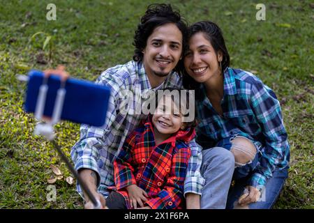 Lateinamerikanische Eltern lächeln mit ihrem 3-jährigen Sohn, während sie im Park ein Foto machen, mit einem Handystick. Familie und Technologiekonzept. Stockfoto