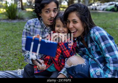 Lateinamerikanische Eltern lächeln mit ihrem 3-jährigen Sohn, während sie im Park ein Foto machen, mit einem Handystick. Familie und Technologiekonzept. Stockfoto