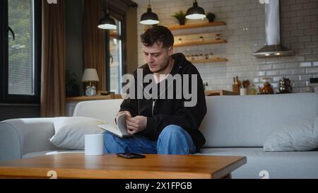 Mann, der auf dem Sofa sitzt, nimmt das Notizbuch vom Tisch, schaut durch seine Seiten, reißt eine Seite ab und wirft das Notizbuch zu Hause auf den Tisch. Stockfoto