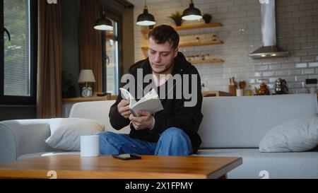 Ein junger Mann, der auf dem Sofa sitzt, nimmt das Notizbuch vom Tisch, schaut durch die Seiten, reißt eine Seite ab und wirft das Notizbuch zu Hause auf den Tisch. Stockfoto