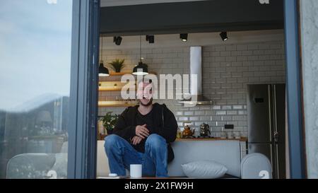 Der überglückte junge Mann trägt Ohrhörer, die auf dem Sofa stehen, singen, im Wohnzimmer tanzen, sieht sorglos aus und hat am Wochenende allein zu Hause Spaß. Stockfoto