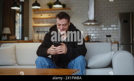 Lächelnder Mann sitzt auf der Couch mit Smartphone Nachricht überprüfen Nachricht lesen Nachricht lesen Empfangen Sie gute Nachrichten per E-Mail Feiern Sie großartige Gelegenheit fühlt sich glücklich Stockfoto