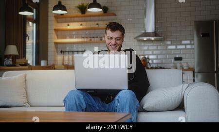 Lächelnder junger Mann, der am Laptop arbeitet, während er auf einem Sofa im Wohnzimmer sitzt. Im Internet surfen, Kaffee aus einer Tasse trinken, zu Hause Spaß haben Stockfoto