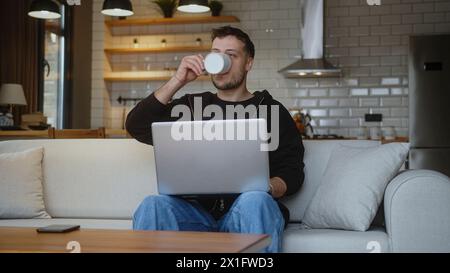 Junger Mann, der am Laptop arbeitet, während er auf einem Sofa im Wohnzimmer sitzt. Im Internet surfen, Kaffee aus einer Tasse trinken, zu Hause Spaß haben. Stockfoto