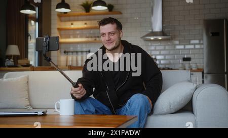 Ein hübscher junger Mann, der auf dem Sofa sitzt, posiert und lächelt, macht Selfie-Fotos mit einem Selfie-Stick im Wohnzimmer zu Hause. Freizeitkonzept Stockfoto