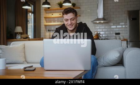 Gutaussehender Mann sitzt auf dem Sofa und führt Videogespräche über einen Laptop zu Hause. Teilen Sie Neuigkeiten, sprechen Sie mit Familie oder Freunden, genießen Sie Webchat Stockfoto