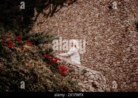 Eine Frau sitzt auf einer Steinmauer mit roten Blumen im Hintergrund. Sie lächelt und sieht in die Kamera. Stockfoto