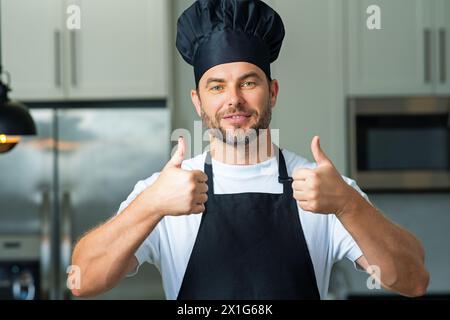 Glücklicher Koch, der mit Daumen nach oben kocht. Porträt eines Küchenmannes in einer Kochmütze in der Küche. Mann mit Schürze und Küchenuniform und Küchenmütze. Zeichen Stockfoto