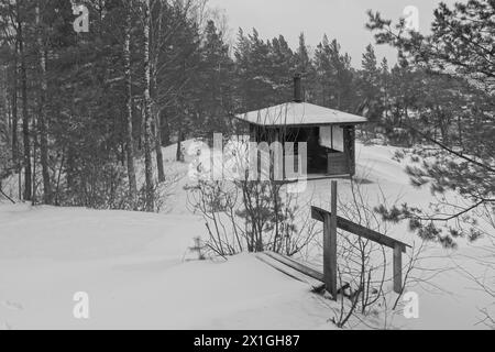 Schneebedeckte Holzkochhütte mit Tisch und Kamin in Vetokannas bei bewölktem Winterwetter, Kopparnäs, Finnland. Stockfoto