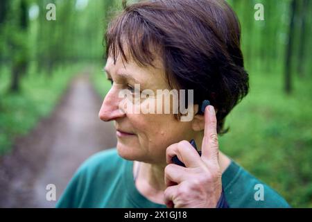 Eine ältere Frau, die im Frühlingswald mit Stöcken Nordic Walking betreibt, schaltet Musik über Kopfhörer Stockfoto