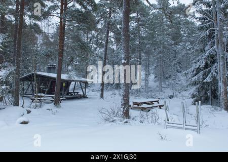 Schneebedeckte Holzkochhütte mit Tisch und Kamin in Vetokannas bei bewölktem Winterwetter, Porkkalanniemi, Kirkkonummi, Finnland. Stockfoto