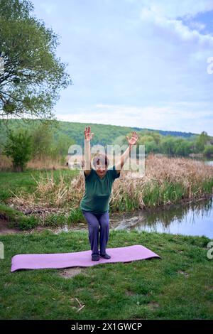 Eine alte Frau praktiziert morgens am Flussufer Yoga, kam von zu Hause mit dem Fahrrad Stockfoto