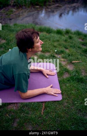 Eine alte Frau praktiziert morgens am Flussufer Yoga, kam von zu Hause mit dem Fahrrad Stockfoto