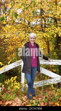 Frau mit schwarzer Jacke und lila Pullover lehnt sich an einen rustikalen weißen Holzzaun. Gelb, Herbstlaub umgibt sie. Stockfoto