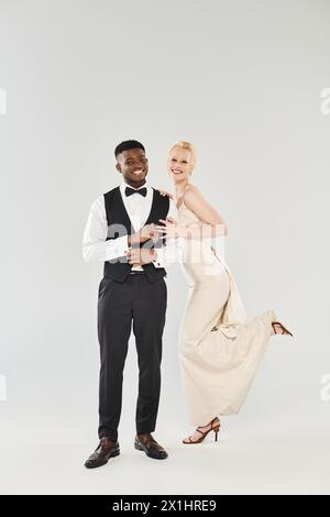 Eine wunderschöne blonde Braut in einem Hochzeitskleid und ein Afroamerikaner im Smoking posieren zusammen in einem Atelier vor grauem Hintergrund. Stockfoto