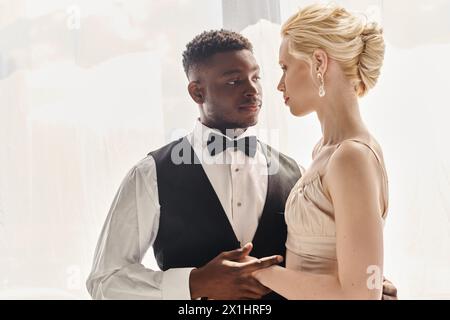 Eine wunderschöne blonde Braut in einem Hochzeitskleid und ein Afroamerikaner, der zusammen in einem Atelier vor grauem Hintergrund steht. Stockfoto