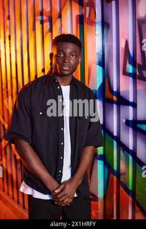 Ein Afroamerikaner steht selbstbewusst vor einer lebendigen, mit Graffiti bedeckten Mauer in einer urbanen Umgebung. Stockfoto