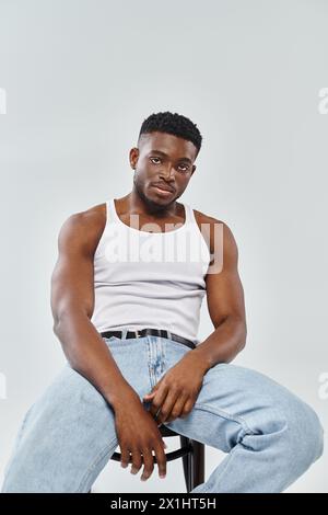 Ein junger Mann eines interrassischen Paares sitzt selbstbewusst auf einem Stuhl, während er Jeans auf grauem Studiohintergrund trägt. Stockfoto