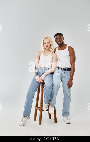 Ein junges, interrassisches Paar sitzt anmutig zusammen auf einem Hocker in einem Studio vor grauem Hintergrund. Stockfoto