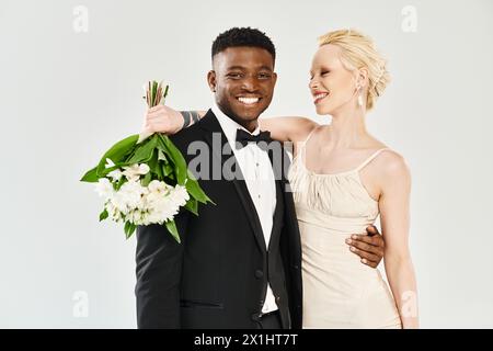 Eine wunderschöne blonde Braut in einem Hochzeitskleid und ein Afroamerikaner im Smoking stehen selbstbewusst zusammen in einem Studio. Stockfoto