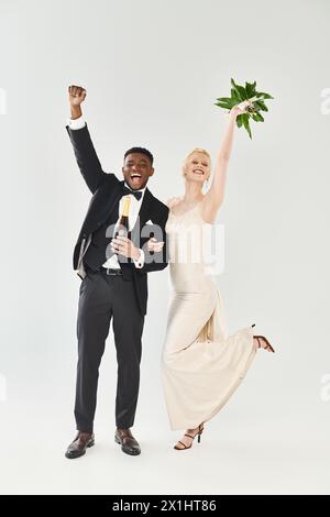 Eine wunderschöne blonde Braut in einem Hochzeitskleid und ein afroamerikanischer Bräutigam, der Blumen in einem Atelier auf grauem Hintergrund hält. Stockfoto