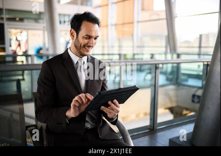 Ein eleganter Geschäftsmann bedient sich eines digitalen Tablets, während er an einem Flughafenterminal auf seinen Flug wartet Stockfoto