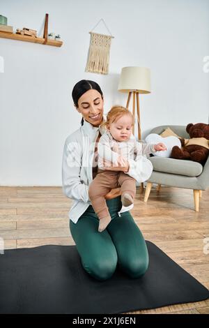 Eine junge Mutter sitzt auf einer Yogamatte und hält ihr Baby friedlich in der Hand, während sie während eines Elternkurses von ihrem Trainer begleitet wird. Stockfoto