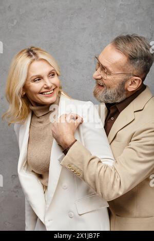 Ein eleganter, reifer Mann hilft einer Frau, ihren Mantel in einer Debonair-Pose vor grauem Hintergrund anzuziehen. Stockfoto