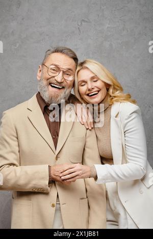 Ein elegantes, reifes, liebevolles Paar in brauner Kleidung, das vor grauem Hintergrund nebeneinander steht. Stockfoto