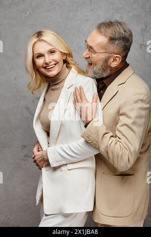 Reife, liebevolle Paare in Debonair-Kleidung stehen anmutig zusammen vor einem grauen Hintergrund. Stockfoto