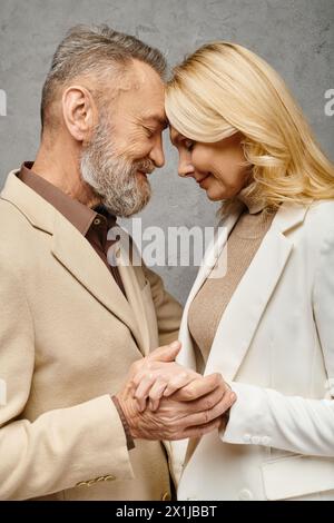 Ein reifes, liebevolles Paar in unbändiger Kleidung, das Hände hält und vor grauem Hintergrund lächelt. Stockfoto