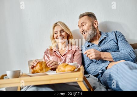 Ein reifes, liebevolles Paar in Hauswäsche, das auf einer Couch sitzt und zusammen ein Tablett Croissants in gemütlicher Umgebung genießt. Stockfoto