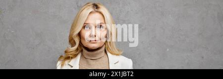 Reife Frau strahlt Eleganz aus in weißer Jacke und Rollkragenpullover vor grauem Hintergrund. Stockfoto