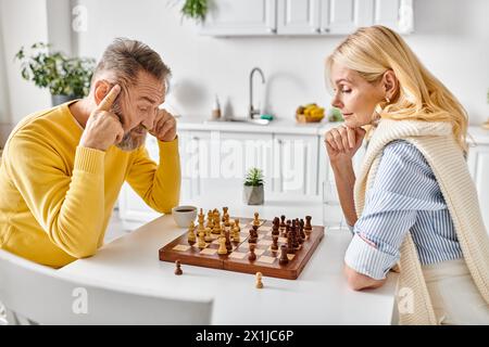 Ein reifes, liebevolles Paar in kuscheligen Hausbekleidung, das sich in einem strategischen Schachkampf an einem Tisch in ihrer Küche zu Hause engagiert. Stockfoto