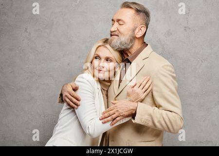 Ein reifes, liebevolles Paar in Debonair-Kleidung umarmen sich in anmutiger Pose vor grauem Hintergrund. Stockfoto