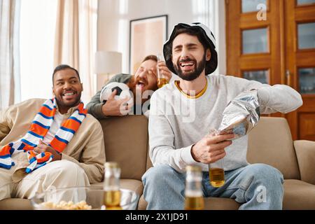 Drei gutaussehende, fröhliche Männer verschiedener Rassen, die sich gegenseitig auf einer Couch in lässiger Kleidung amüsieren. Stockfoto