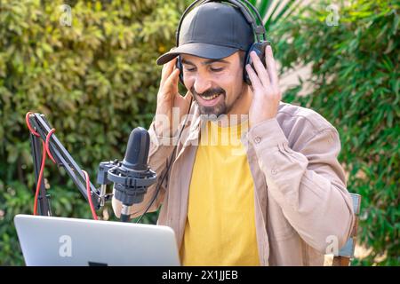 Ein Mann mit Kopfhörern, der Podcasts in ein Mikrofon aufnimmt, während er im Freien einen Laptop benutzt Stockfoto