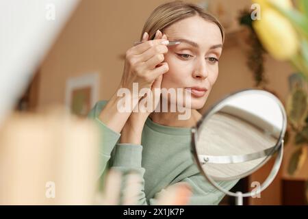 Warm getöntes Porträt einer eleganten erwachsenen Frau, die Augenbrauen mit einer Pinzette zupft und im Spiegel im Kopierraum zu Hause schaut Stockfoto