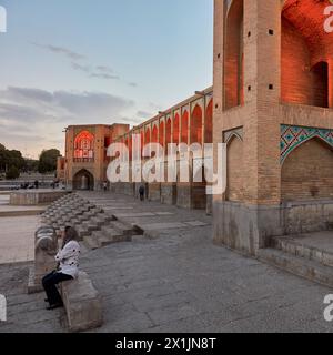 Iranische Frau sitzt auf einer Steinbank im beleuchteten 17. Jahrhundert. Khaju-Brücke auf dem Fluss Zayanderud während der Trockenzeit mit trockenem Flussbett. Isfahan, Iran Stockfoto